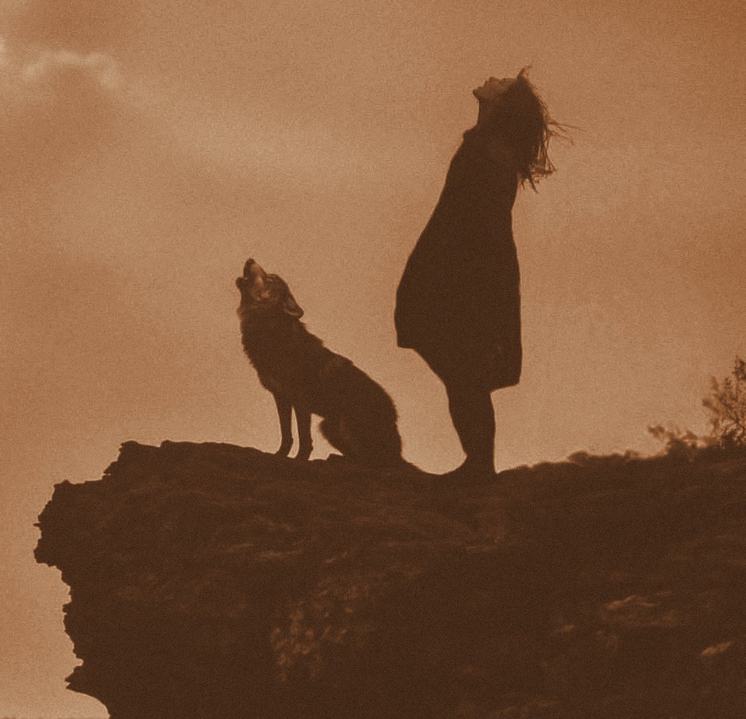 Silhueta de uma mulher e de um lobo no alto de uma rocha, os dois de cabeça erguida, uivando para o céu