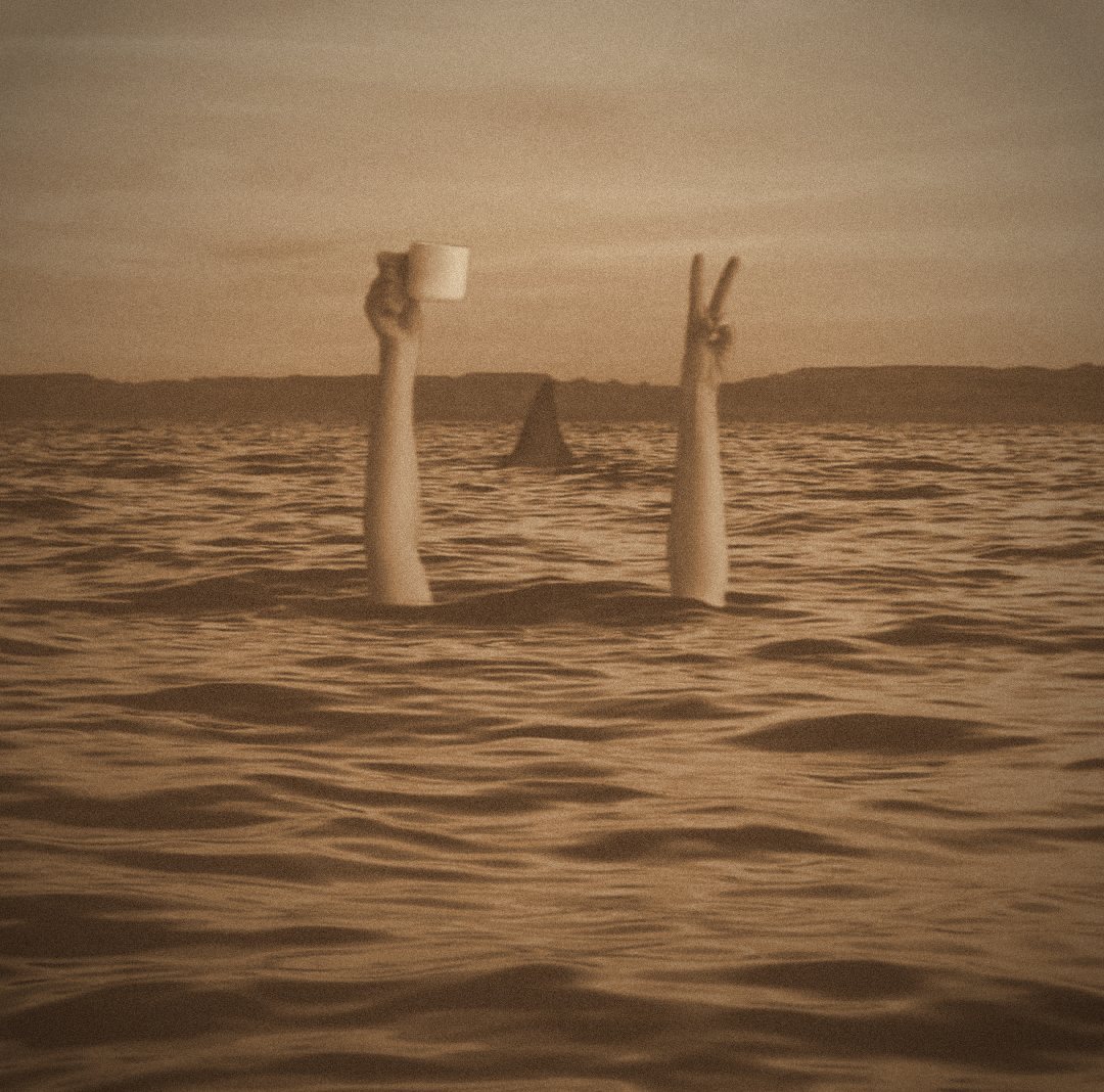 Foto em sépia: dois braços saem do mar, um segura uma xícara de café e o outro faz o sinal de paz, com uma barbatana de tubarão ao fundo